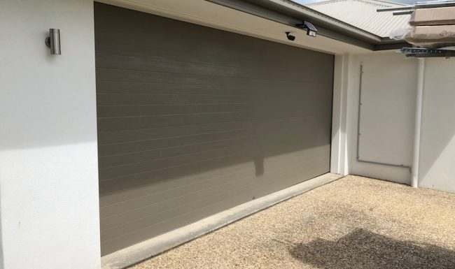 Grey-brown sectional garage door closed against a rendered wall beside an outdoor light